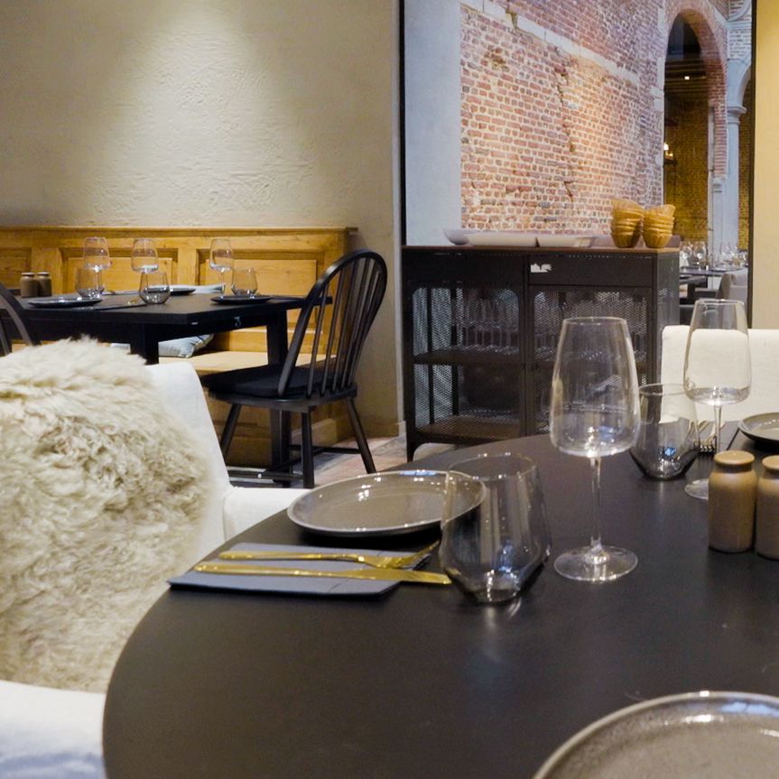 Stijlvol restaurantinterieur met zwarte ronde tafel, glazen, bestek en kaars. Zicht op bakstenen muur en boog via grote glazen wand.