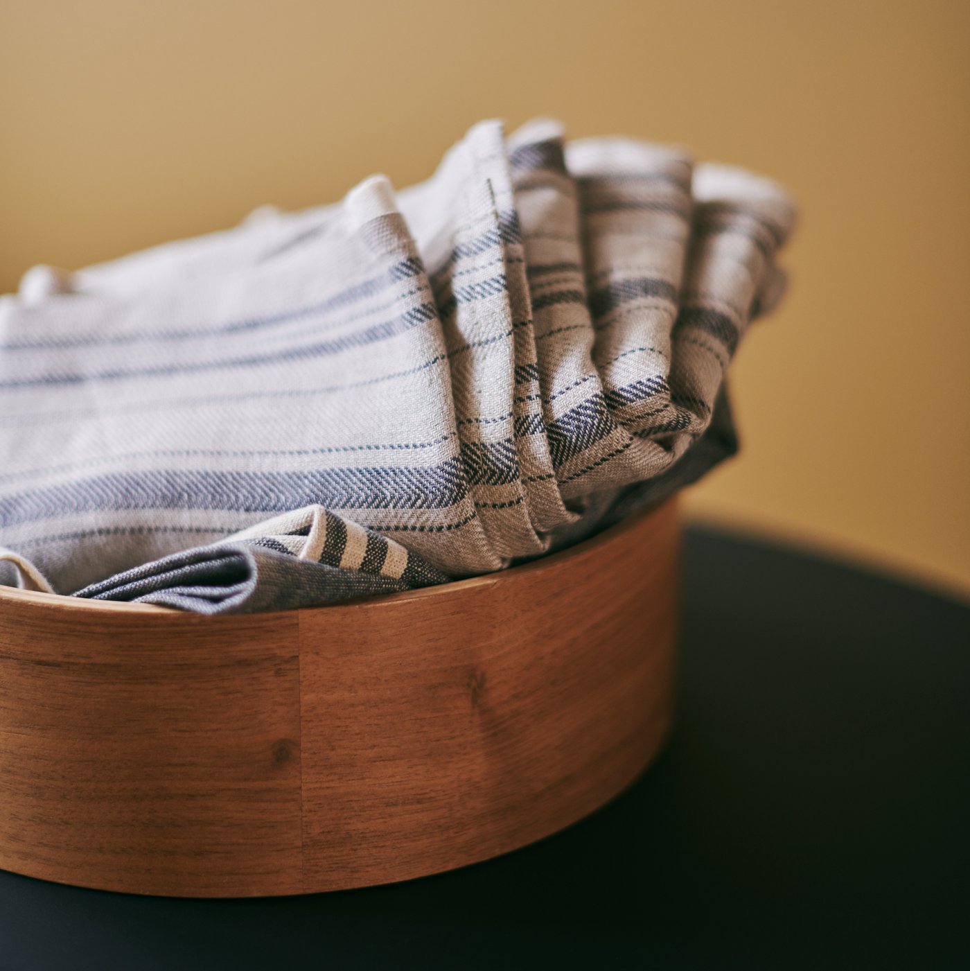 Cotton tea towels are neatly folded and stacked in a wooden bowl.