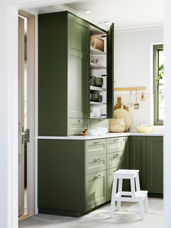 A kitchen featuring AXSTAD grey-green fronts with an open tall cupboard, white countertops, and a small white step stool.