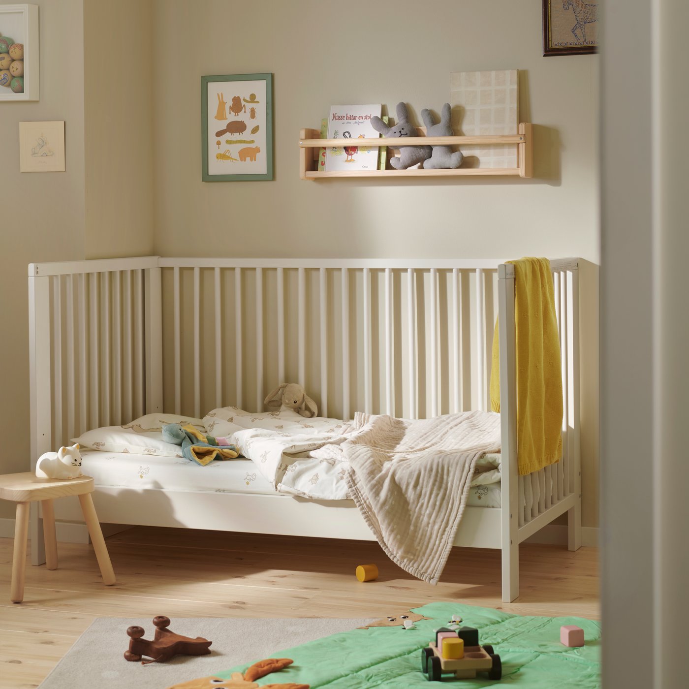 A GULLIVER cot in white wood in a neutral nursery, with bedding and a yellow towel, and toys on a green play mat below.