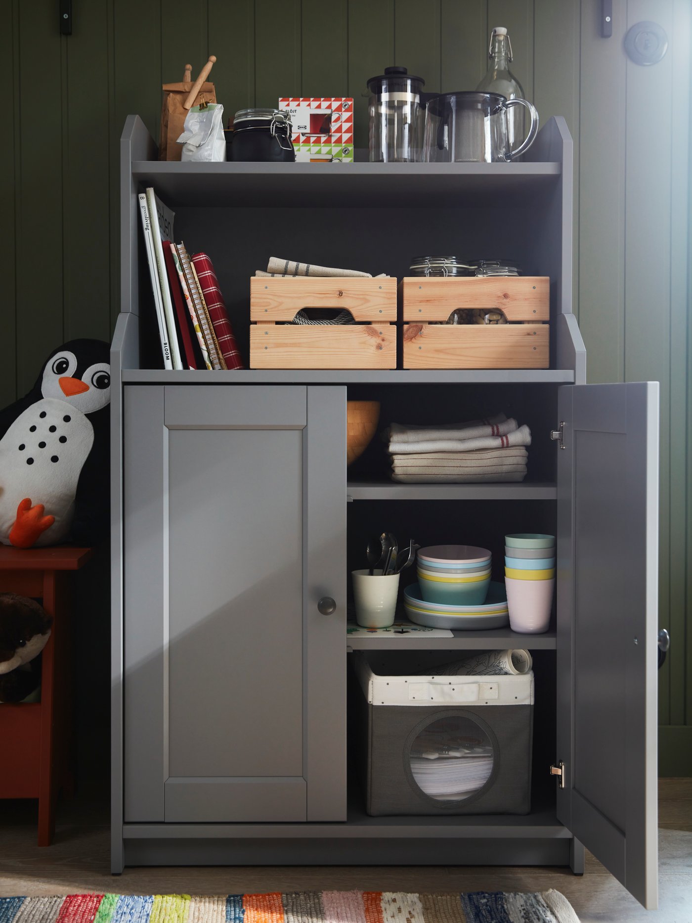 A grey HAUGA cabinet with 2 doors holding KALAS tableware, a LEN box and two pine KNAGGLIG boxes stands against a wall.
