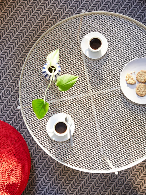 An aerial view of a beige, circular KROKHOLMEN outdoor coffee table with coffee cups, a plant and a plate of cookies on top.