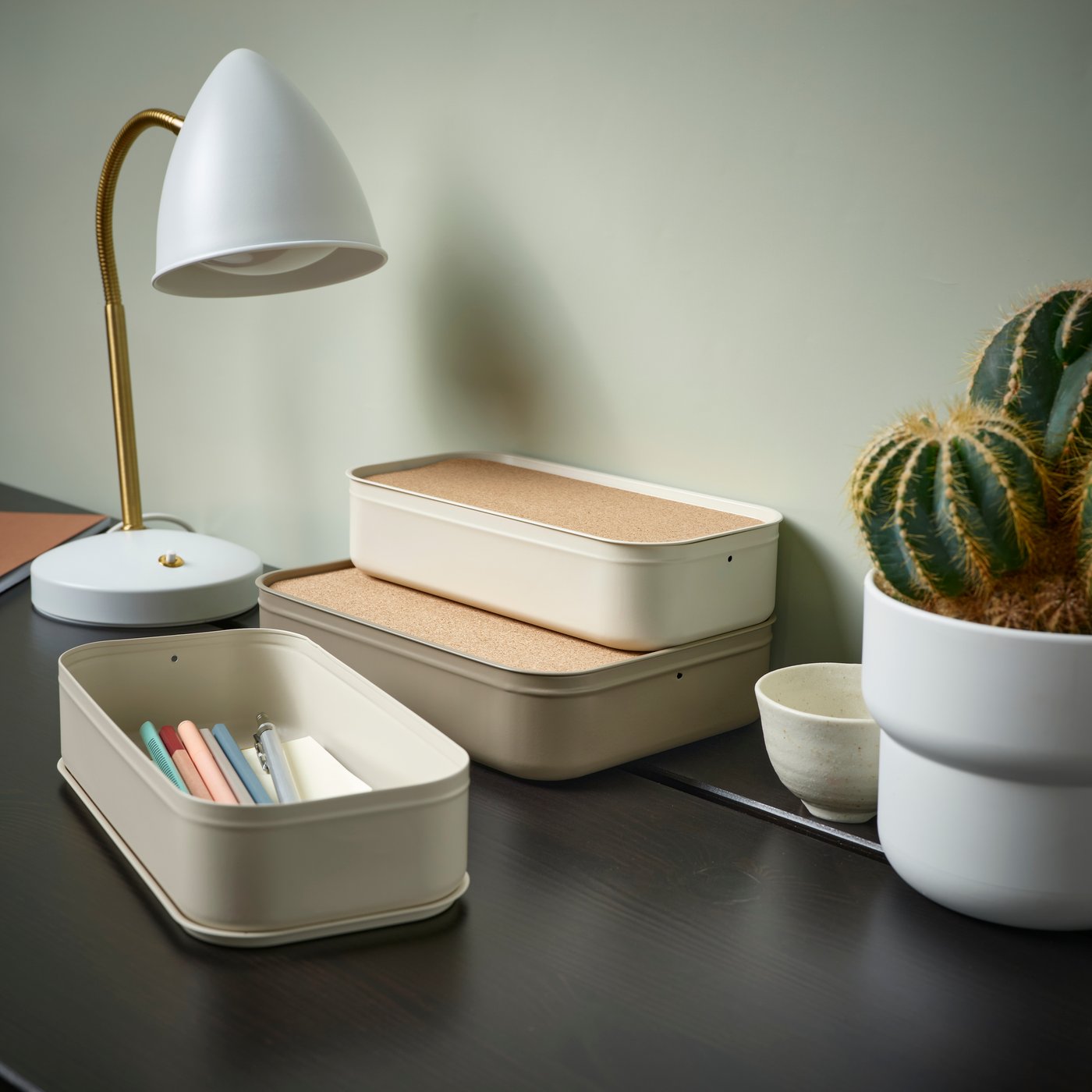 Desk setup with a white lamp, stacked beige storage boxes, pencils in a tray, and a potted cactus.