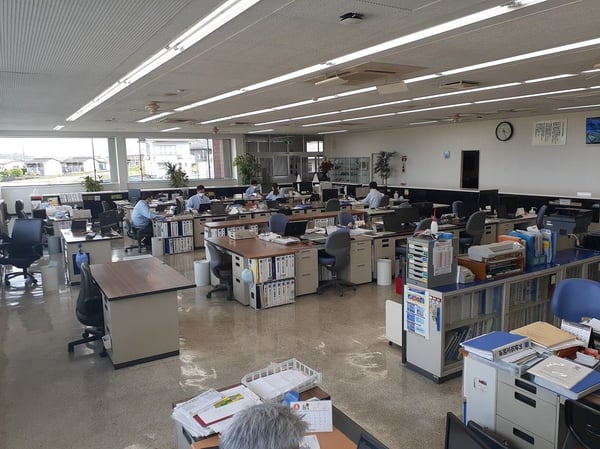 デスクと書類棚が並ぶ広いオフィスフロア。天井の蛍光灯と大きな窓からの光で明るく照らされた執務スペース。