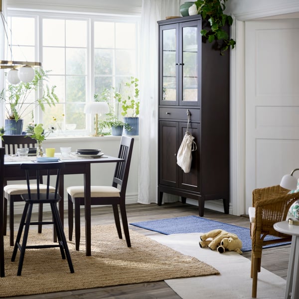 A dark brown NÄSINGE chair with light beige cushions stands by a sunlit window in a bright dining area filled with greenery.