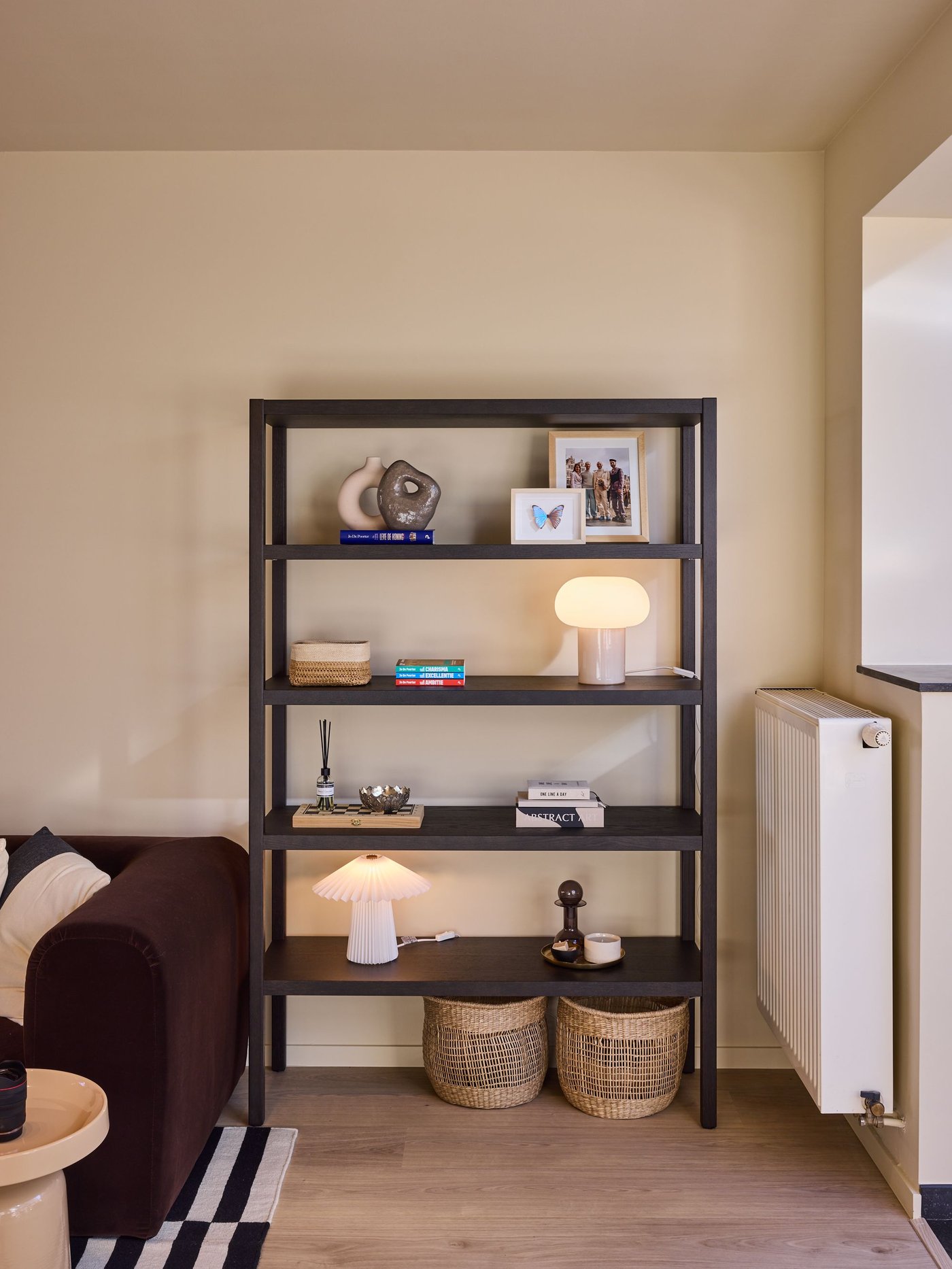 Zwarte open rek met decoratie, lampjes en manden tegen een beige muur, naast radiator en zetel in een warme woonruimte.