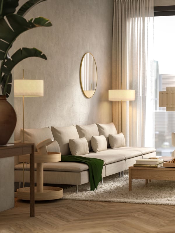I ett modernt lägenhets vardagsrum med stora fönster finns en lång, beige SÖDERHAMN modulsoffa och två soffbord i ekfanér.