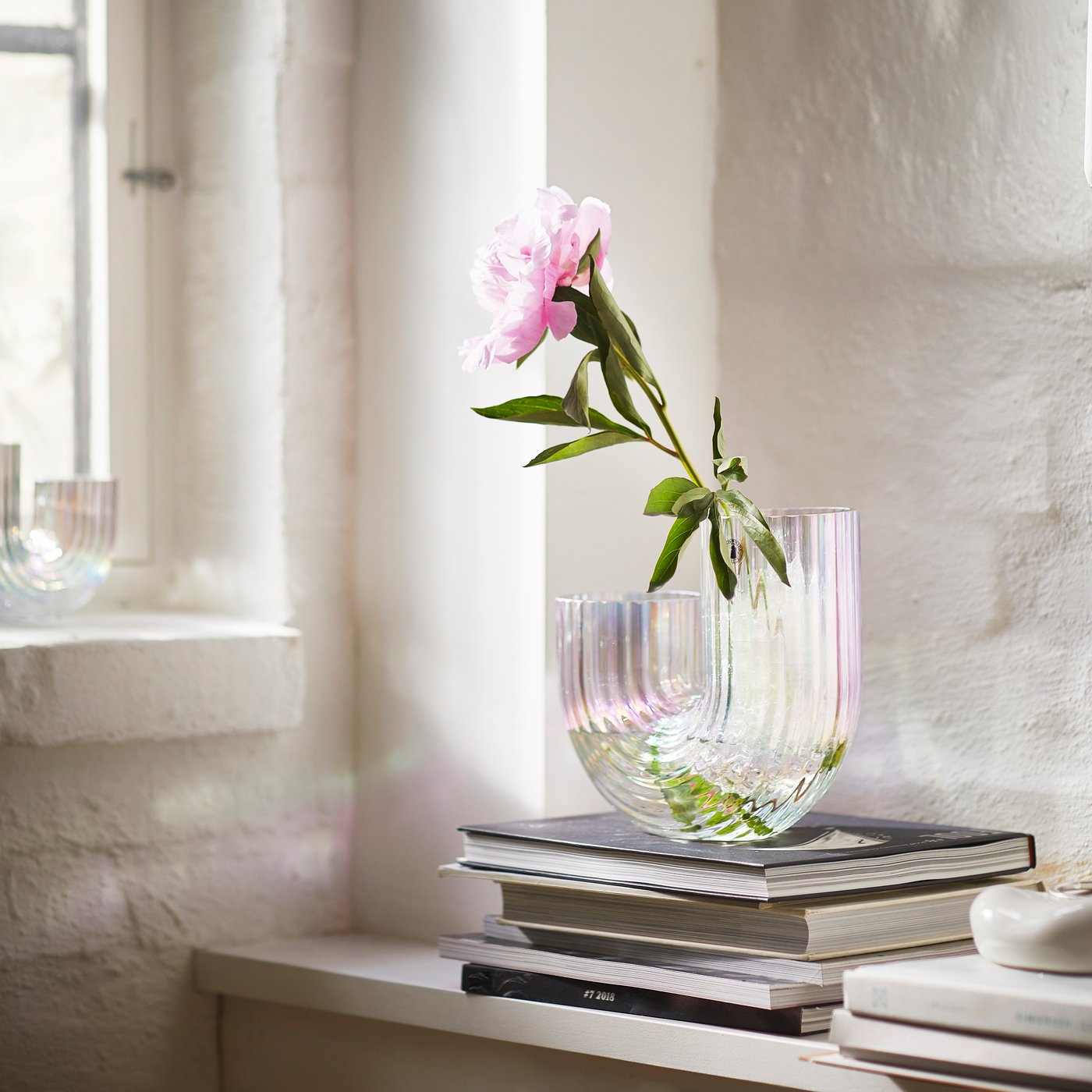 Vaso RÄFFELBJÖRK color madreperla con un fiore rosa, appoggiato su una pila di libri vicino alla finestra per creare un angolo luminoso e delicato.