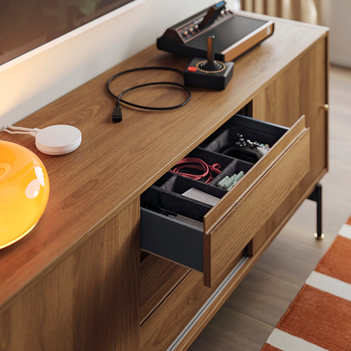 An open drawer of a RÅDMANSÖ TV bench contains filled RAGGISAR trays. A DIRIGERA hub and gaming console are on the bench.