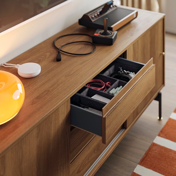 An open drawer of a RÅDMANSÖ TV bench contains filled RAGGISAR trays. A DIRIGERA hub and gaming console are on the bench.