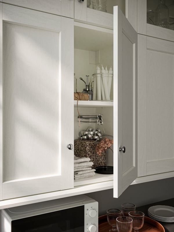Une armoire de cuisine ouverte avec des portes ENKÖPING blanches, sur les tablettes se trouvent des bougies, des serviettes et un bocal KORKEN avec couvercle.