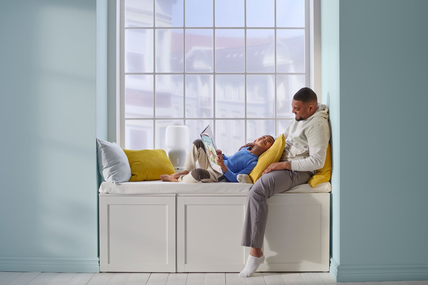 Two people sitting on a bench seat with lots of cushions next to a large window