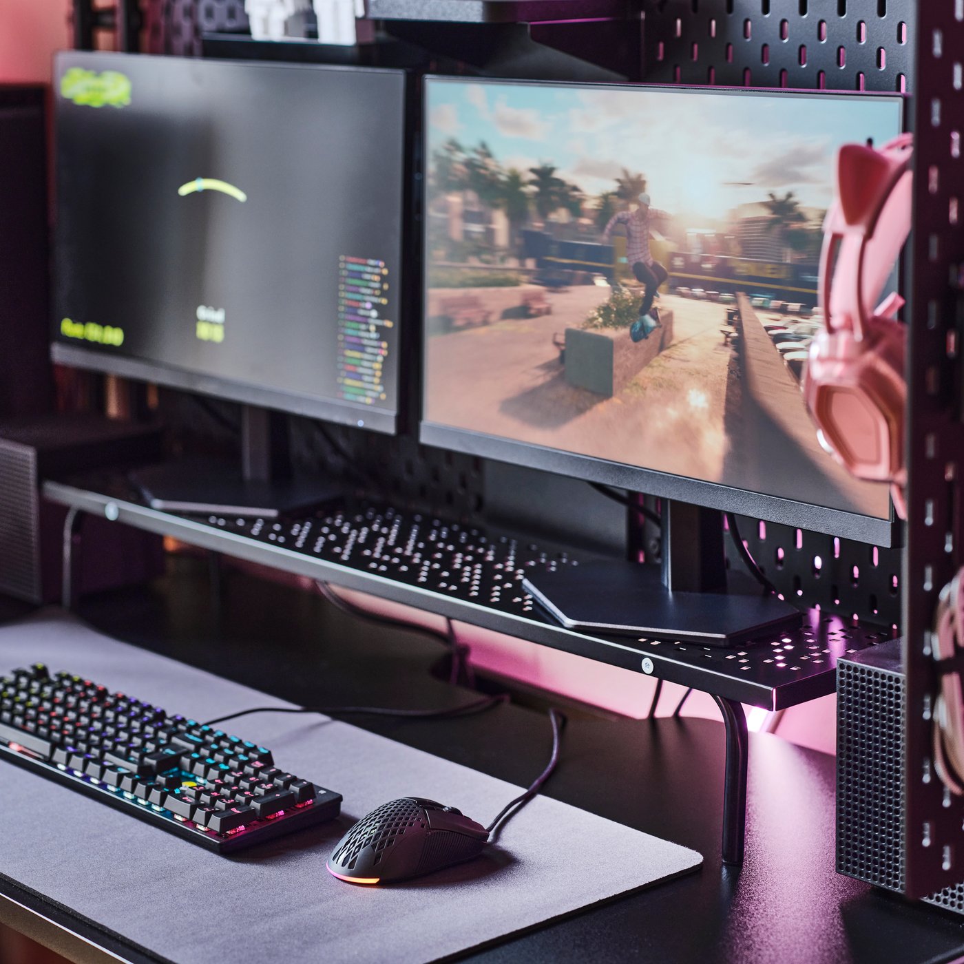 Una estación de juego con un soporte para monitor LÅNESPELARE negro colocado debajo de dos pantallas, con un teclado y un mouse cerca.