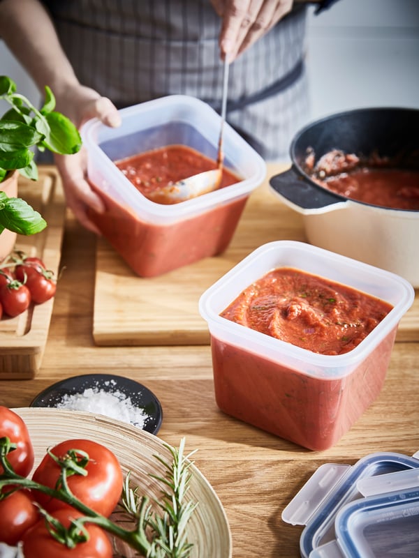 A person wearing a checked apron is spooning tomato sauce from a pan into two plastic food containers.
