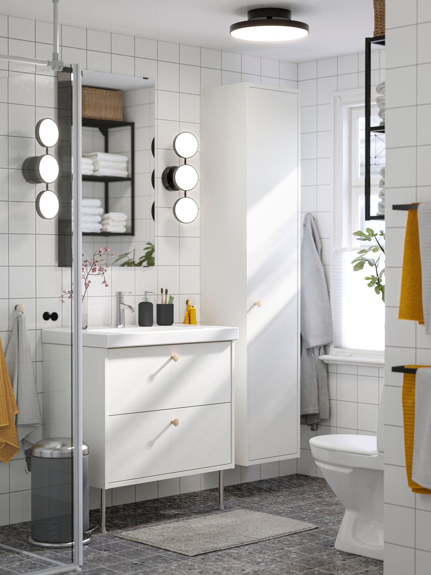 A white-tiled bathroom has a white HAVBÄCK wash-stand with drawers, two black KABOMBA LED wall lamps and a grey bathroom mat.