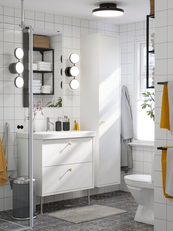 A white-tiled bathroom has a white HAVBÄCK wash-stand with drawers, two black KABOMBA LED wall lamps and a grey bathroom mat.