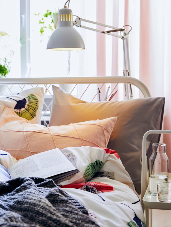 Une lampe de travail blanche TERTIAL au-dessus de la tête d’un lit NESTTUN avec des oreillers, des coussins et une couette colorée.