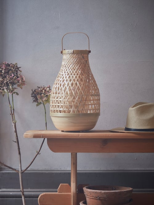 Une lampe de table MISTERHULT en bambou tissée en forme d’ampoule avec une poignée est posée sur un banc en bois, à côté d’un chapeau et de fleurs séchées.