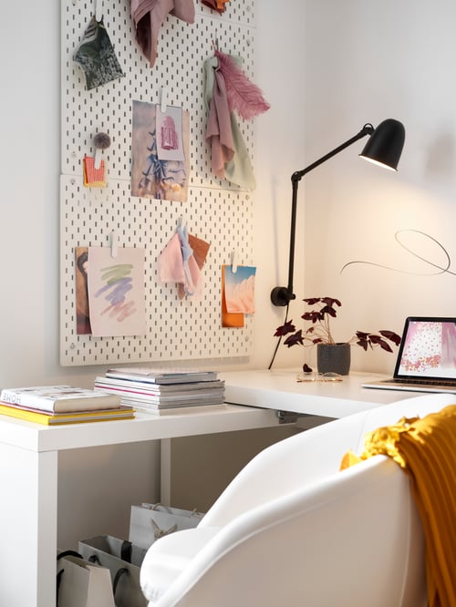A white MALM desk with pull-out panel is in a corner by a white chair. A laptop and stacked magazines are on the desk.