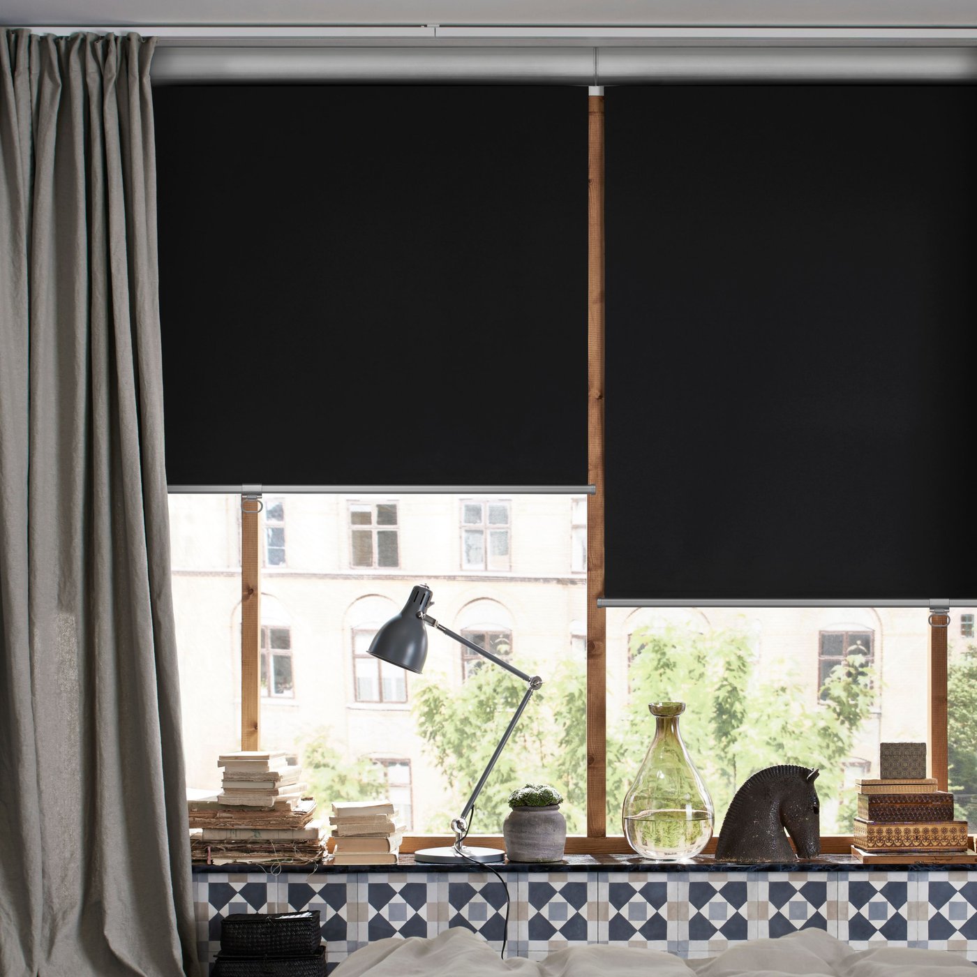 Due tende oscuranti TRETUR grigio scuro sui vetri di un'ampia finestra e una lampada da lavoro appoggiata sul davanzale in una camera da letto beige.