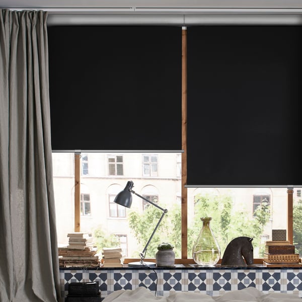 Two dark grey TRETUR block-out blinds on a large window at the end of a beige bedroom with a work lamp on the window sill.