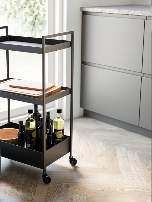 Modern kitchen featuring a black trolley holding different kitchen plates and bottles next to a base cabinet with three drawers. 