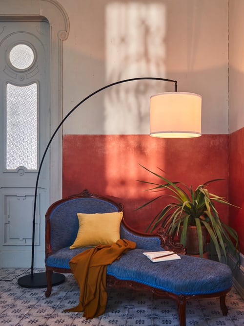 A black metal SKAFTET arched floor lamp base with a white lampshade stands in the corner of a room, by an old chaise longue.
