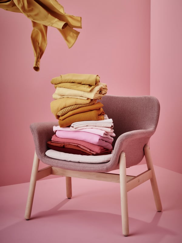 Stapel med vikta kläder i gult, rosa och vitt på en rosa VEDBO stol med träben mot rosa bakgrund.