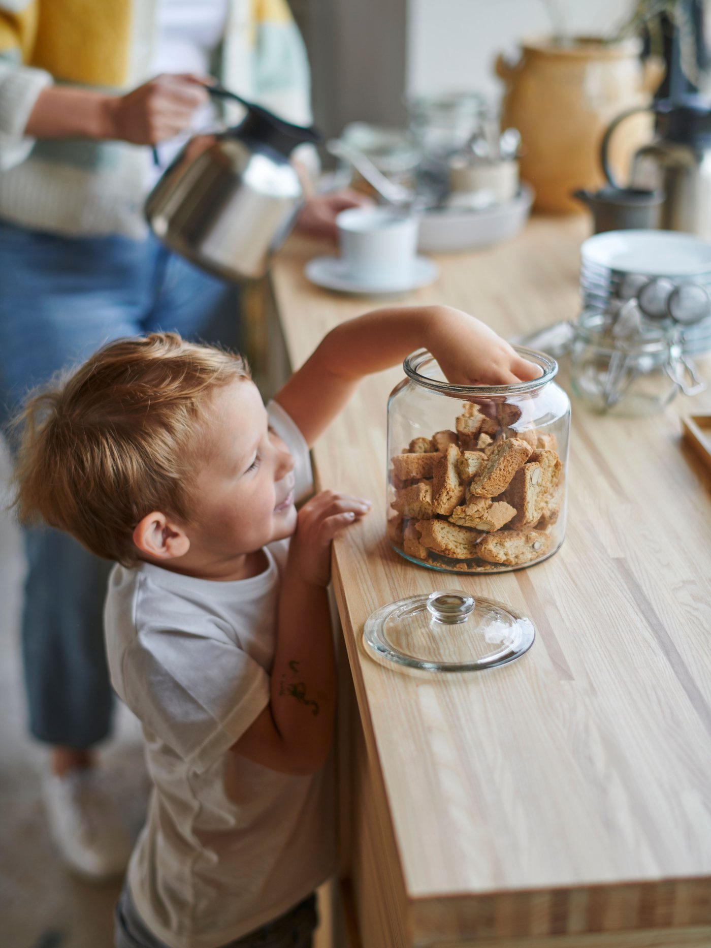 A child is reaching into a VARDAGEN jar filled with crusts. A person is pouring coffee in to cup behind the child.
