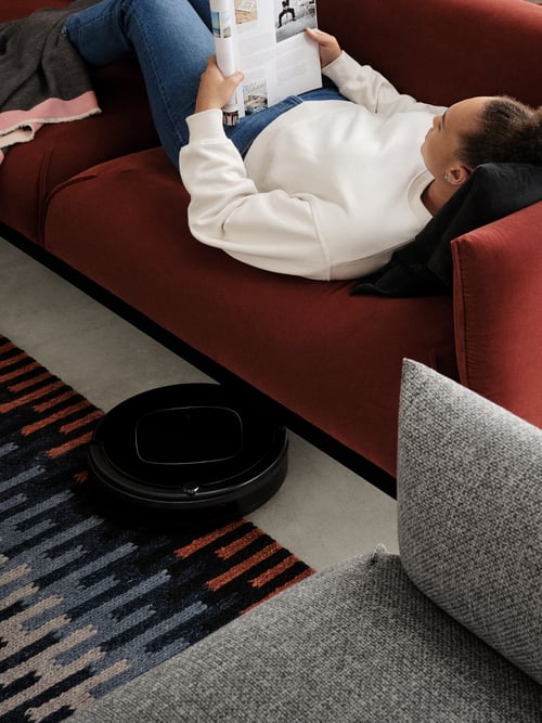 A woman lying on a dark red ÄPPLARYD sofa with her head on a black cushion, reading. A robot vacuum cleaner is moving under the sofa.