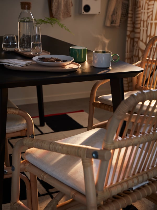 Steaming PIGGÅL mugs on a sun-dappled black LISABO table flanked by SALNÖ rattan chairs, all standing on a RASTPLATS rug.