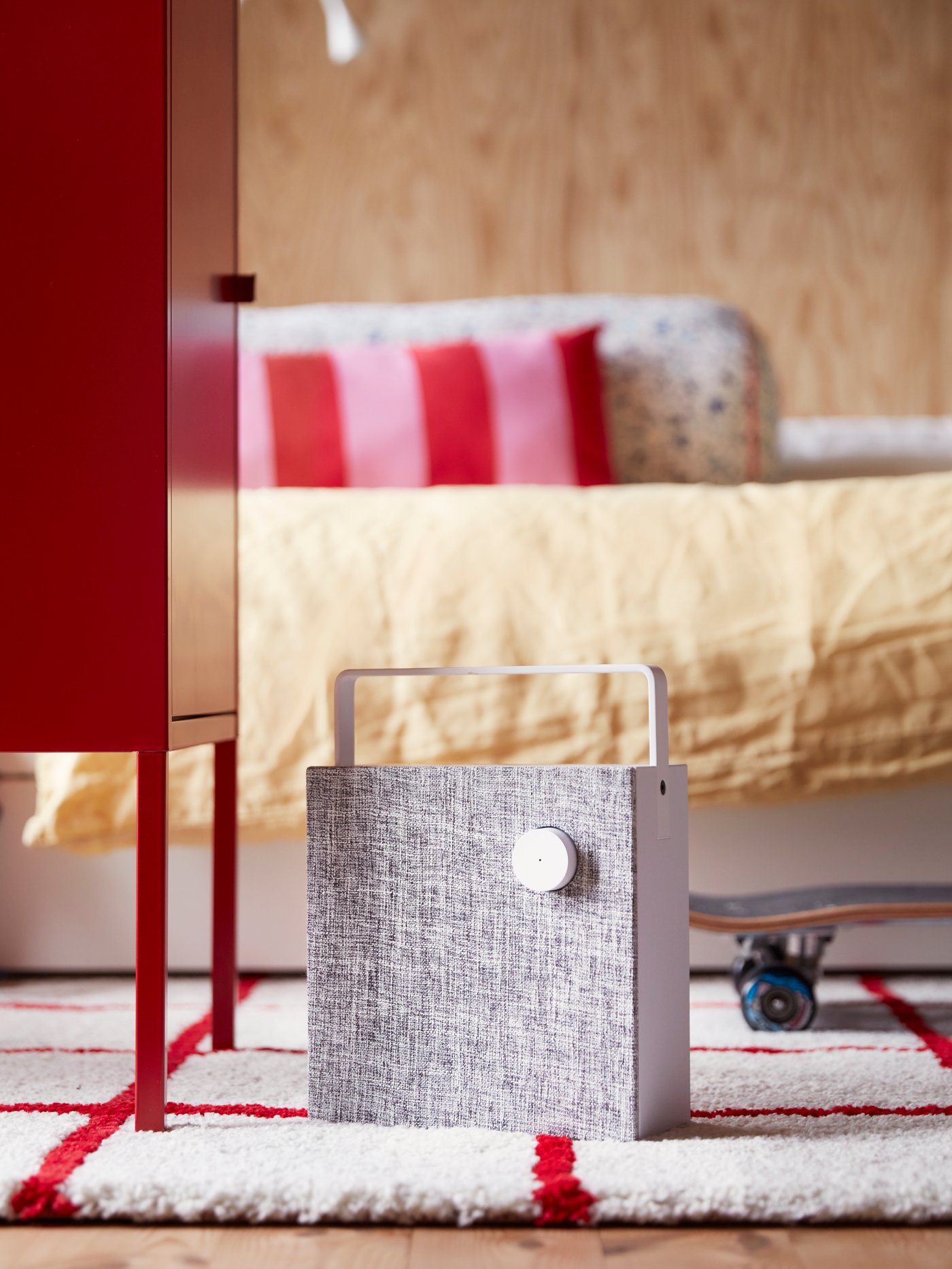 Ein Teenagerzimmer mit dem tragbaren ENEBY Bluetooth®-Lautsprecher auf einem rot-weiß gemusterten Teppich vor einem Bett mit gelber Bettwäsche.