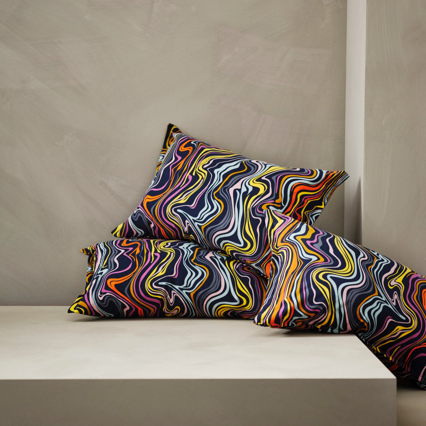 Tiga sarung kusyen KLIPPNEJLIKA berwarna biru gelap/beraneka warna terletak di penjuru platform di ruang studio berwarna kuning air.