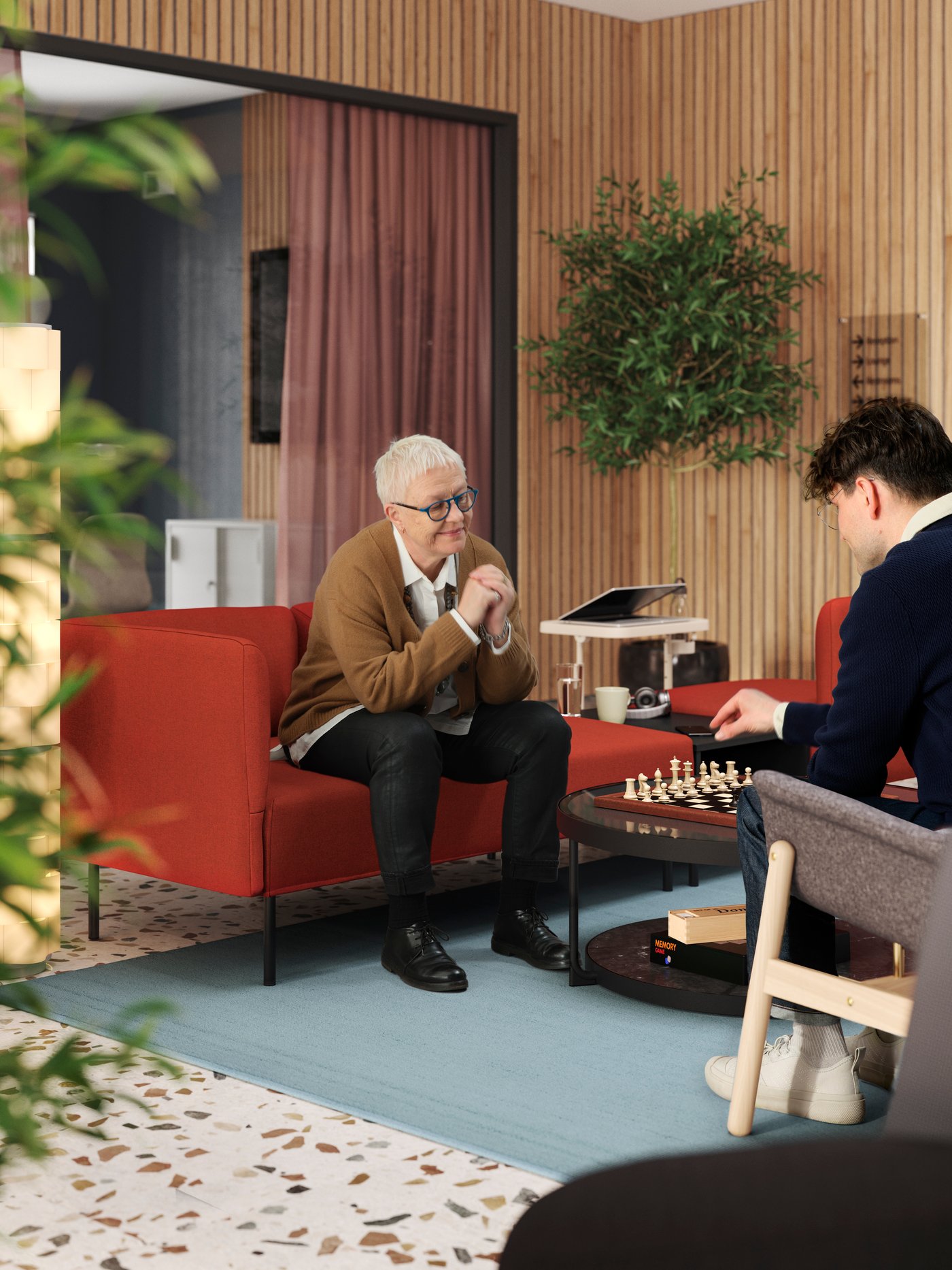 In a modern, wood-panelled lobby is a red LILLEHEM modular sofa with a person sitting on it, waiting on their chess opponent.