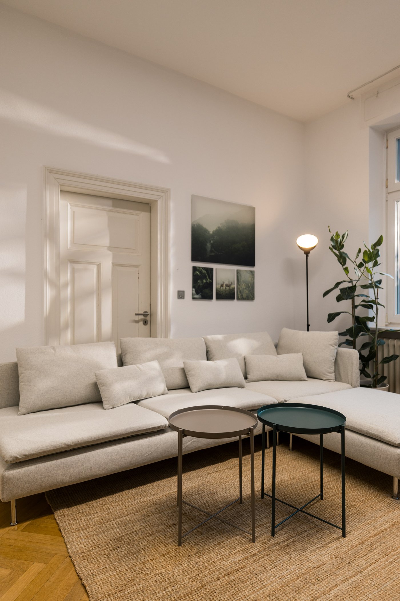 Heller Wohnbereich mit großem Ecksofa, runden Beistelltischen, Stehlampe und Wandbildern.