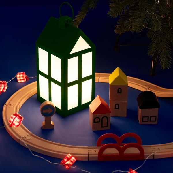 Liten huslykta omgiven av ett LILLABO trätågsspår med färgglada småhus, juldekorationer och ljusslinga – en mysig och festlig julscen.