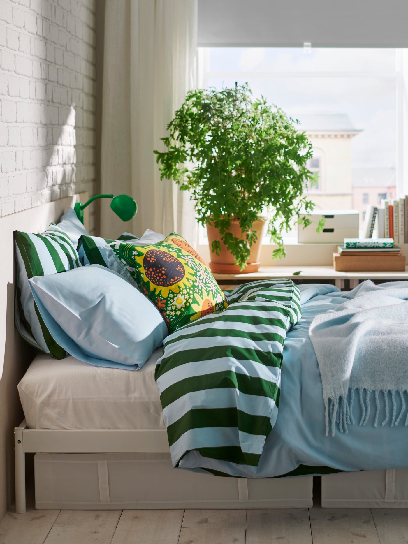 Une chambre lumineuse avec une fenêtre, des plantes et un lit blanc habillé d'une parure de lit SLÅNHÖSTMAL à rayures bleues et vertes.
