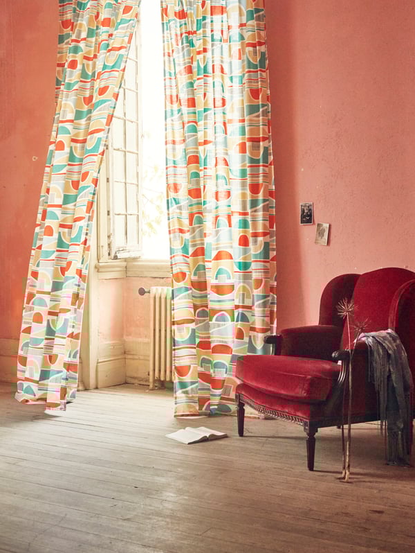 MARTORN curtains with a multi-coloured graphic pattern blowing in the wind of an open window next to an armchair.