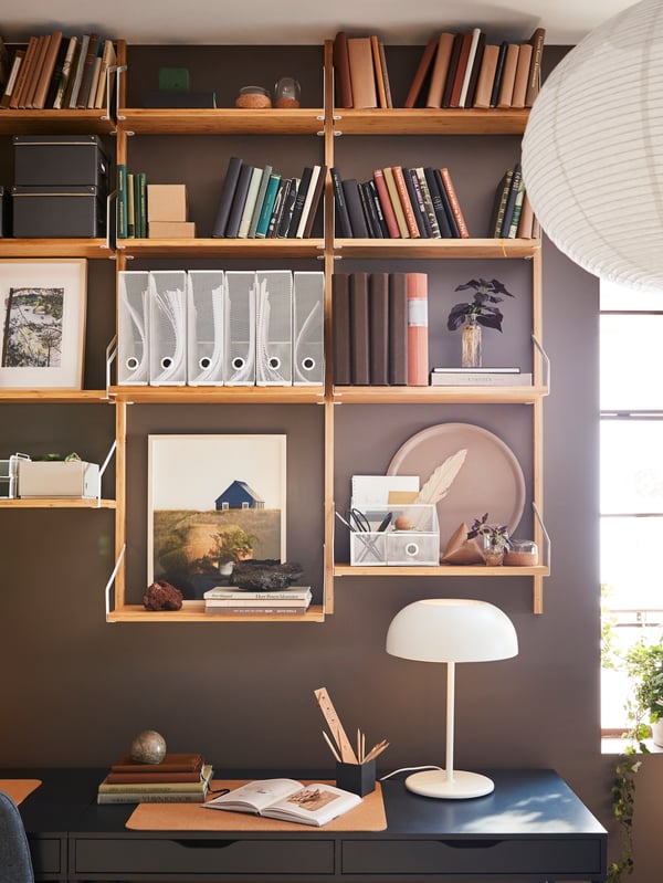 A brown-painted wall with a wall-mounted shelf combination in bamboo with books, decorative items and white files.