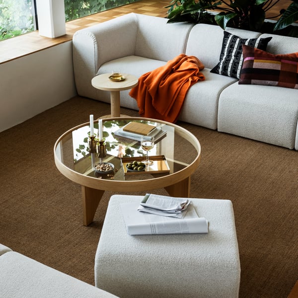 A modern living room with beige sofas topped with an orange throw, a wooden coffee table with a glass top, and a pouffe.