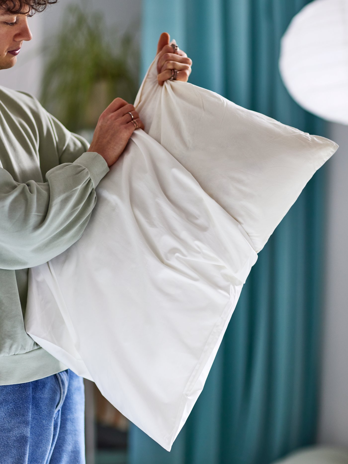 A man putting a white pillowcase on a pillow with a LUDDROS pillow protector, with a plant, curtain and pendant behind.