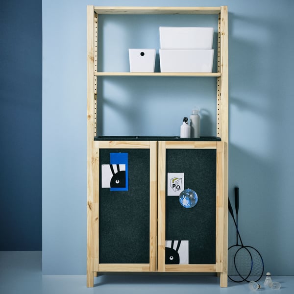 An IVAR shelving unit with two doors, with three white KUGGIS boxes with lids on a shelf, stands against a blue wall.