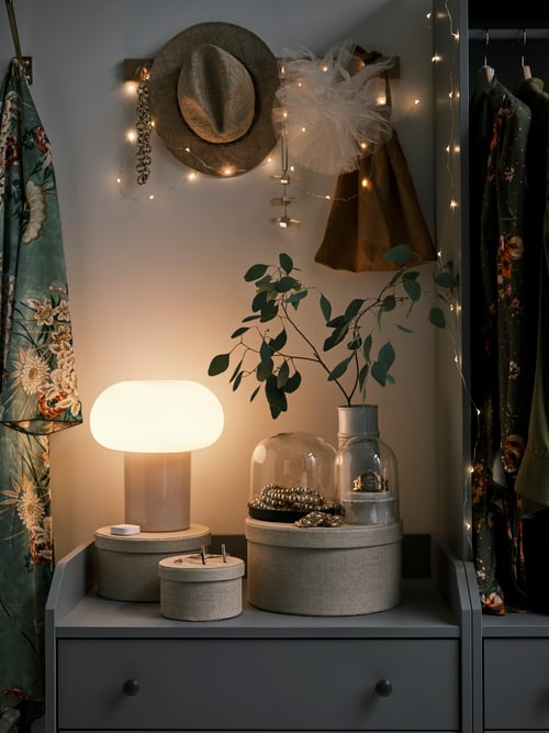 Dimly lit bedroom featuring a lit table lamp placed net to round storage boxes. A lit lighting chain hangs above the lamp.