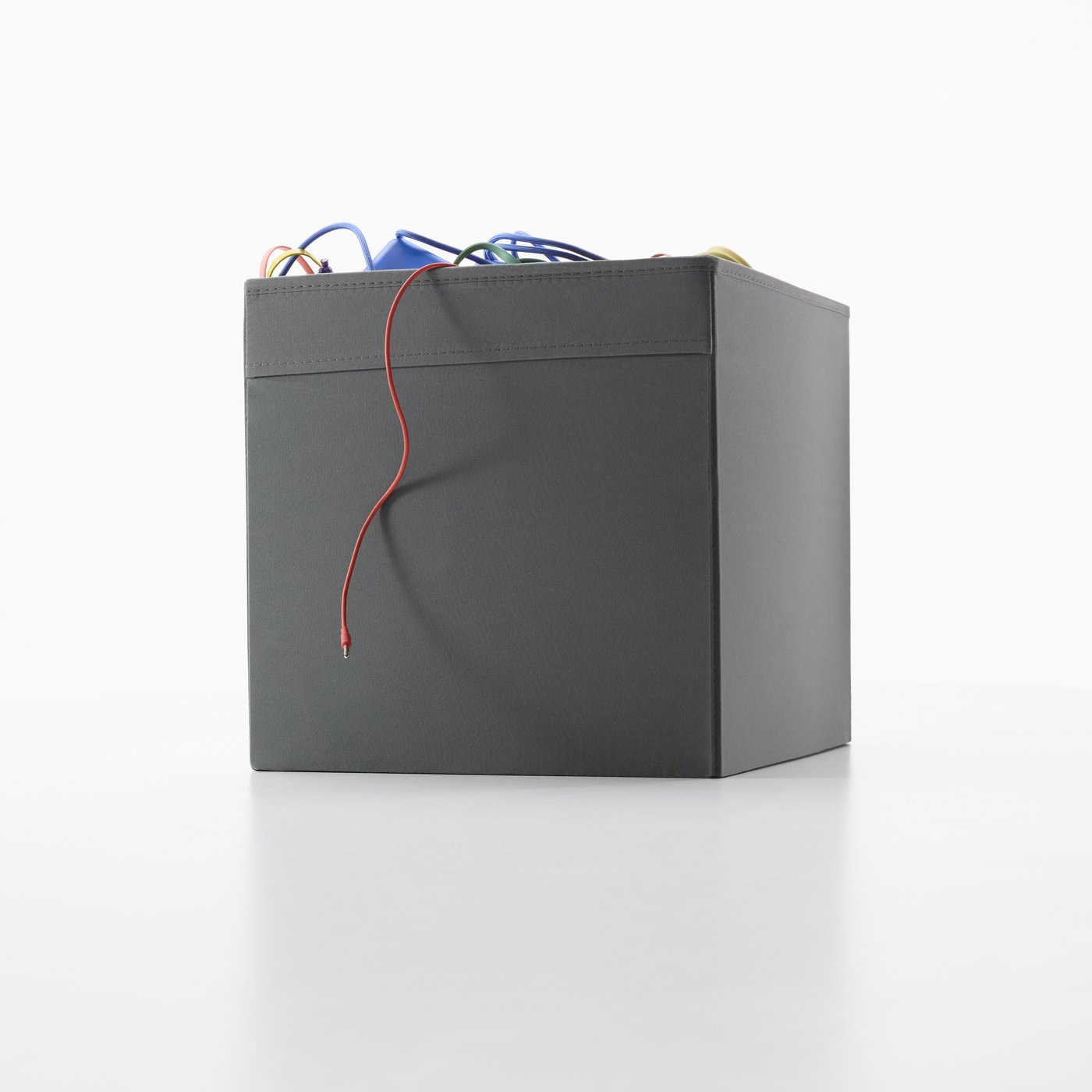 A dark grey DRÖNA box is standing on the floor. Some colourful cables are sticking out.