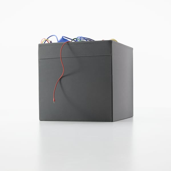 A dark grey DRÖNA box is standing on the floor. Some colourful cables are sticking out.