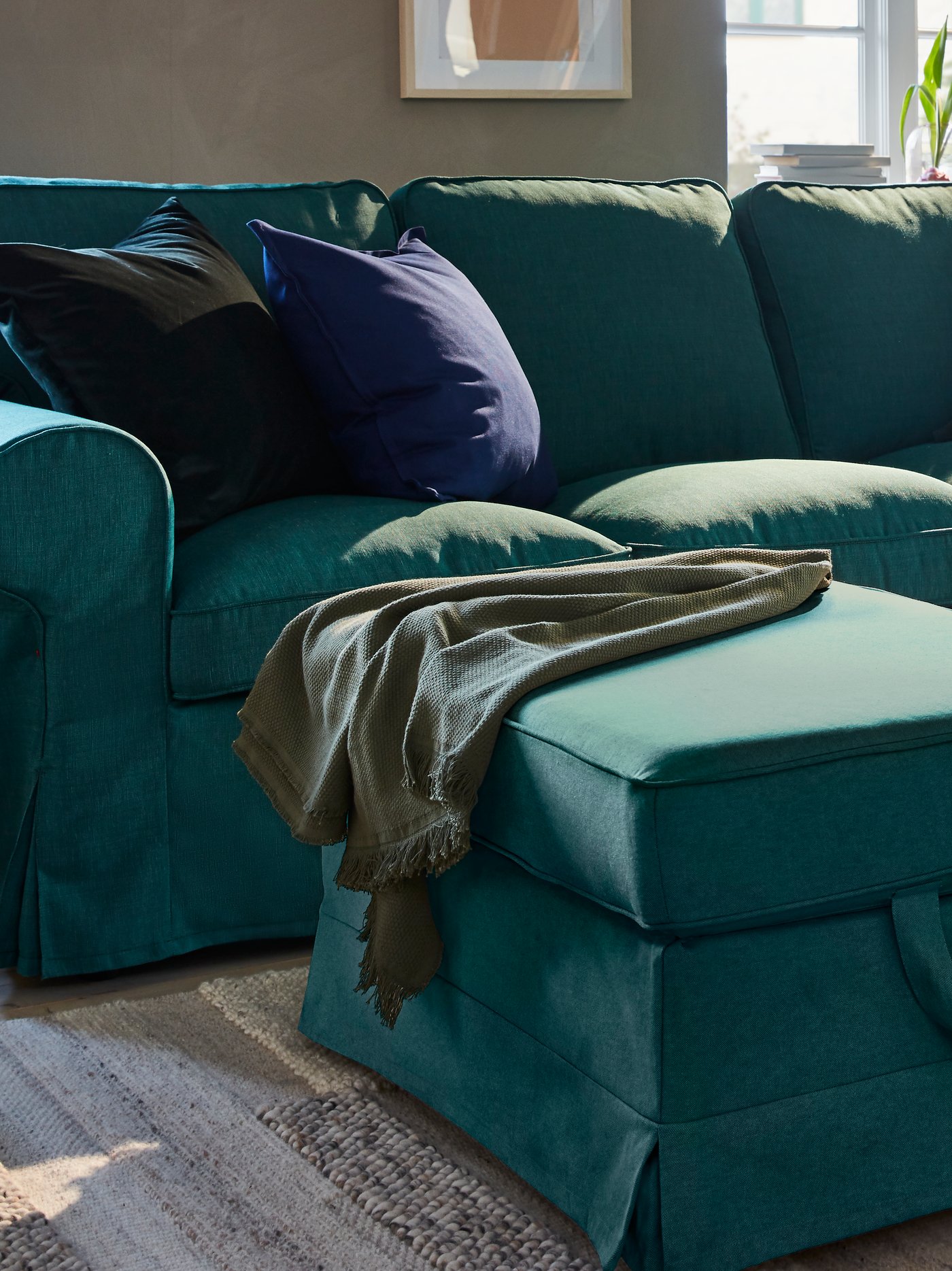 غرفة جلوس بها صوفا لون فيروزي داكن ومسندم قدم مع وسائد وبطانية على سجادة منسوجة يدويًا باللون الأبيض العاجي.