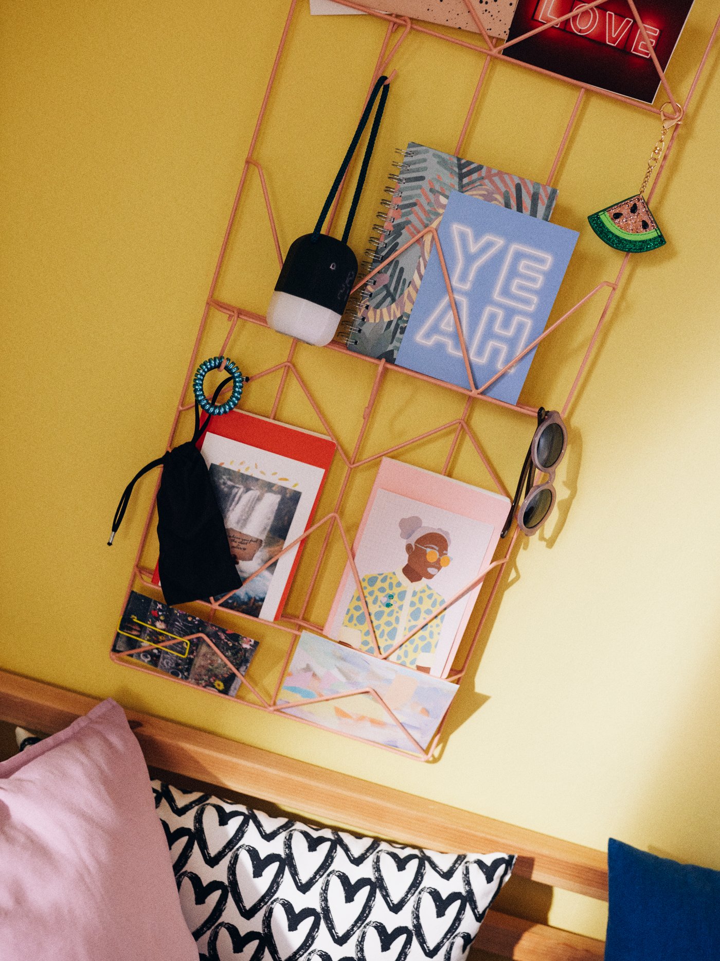 A KVICKSUND memo board on a yellow wall holding cards, sunglasses, notebooks, pictures and keyrings, above colored cushions.