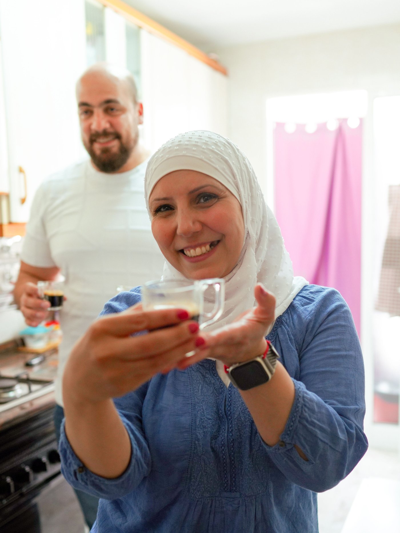 Zahra und Tarek stehen in ihrer Küche und lächeln in die Kamera, während sie Kaffeetassen halten.