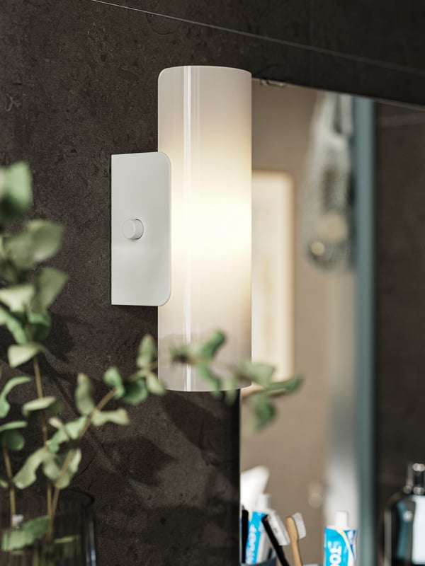 A white HAVSDUN LED wall lamp with frosted glass is dimly lit and hangs next to a mirror on a dark brown wall.