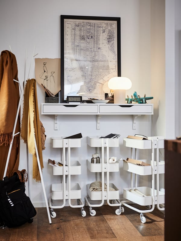 Three white RÅSKOG trolleys next to a white EKRAR hat and coat stand, below a wall shelf with drawers, a map on the wall.